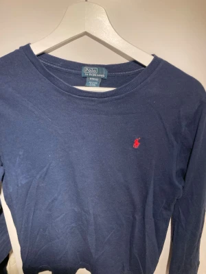 Mörkblå långärmad topp Polo Ralph Lauren - Säljer en mörkblå långärmad topp från Polo Ralph Lauren. Toppen har en klassisk rund halsringning och är dekorerad med den ikoniska röda broderade loggan på bröstet. Tillverkad i mjuk bomull som känns skön mot huden.