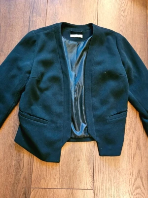 Grön kavaj från Vila - Stilren grön kavaj från Vila med öppen front och två fickor framtill. Klassisk rak passform och helfodrad insida. Perfekt för att lyfta vilken outfit som helst med en clean och enkel look.