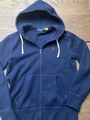 Ralph lauren tröja - Hej jag säljer en Ralph Lauren hoodie då den var förliten när jag köpte den. Med  modern stil Mörkblå Ralph Lauren-tröja i mycket fint skick. Full zip framtill, vita snören på var sin sida  och med en röd logga som ger en stilren men sportig look. Modern passform som funkar perfekt till både vardag och streetwear. Tidlös och lättmatchad.och 100% äkta!