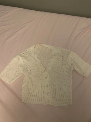 Vit stickad kofta med pärlknappar - Supersöt vit stickad kofta med kort ärm och v-ringad hals. Koftan har fina pärlknappar framtill och ett luftigt, hålmönstrat material som känns perfekt till våren eller sommaren. Croppad modell som ger en trendig look.