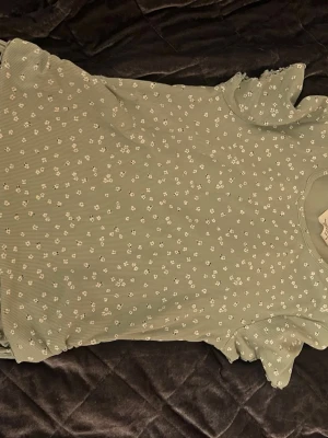 Grön blommig topp från H&M - Söt grön topp från H&M i storlek 146-152. Toppen har  små vita blommor över hela tyget. Toppen har korta ärmar med lätt volangkant och rund halsringning. Materialet är ribbat och stretchigt. Funderingar? Skriv till mig💕