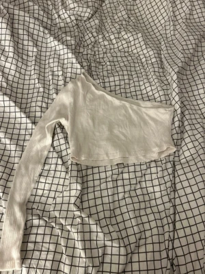 Vit ribbad oneshoulder topp - Säljer en vit ribbad oneshoulder topp med lång ärm på ena sidan. Toppen är croppad och har en tight passform som framhäver axlarna. Perfekt för dig som gillar minimalistisk stil och vill ha något trendigt och enkelt.
