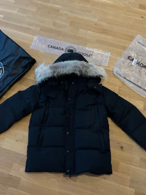 Svart Canada Goose dunjacka med päls - Säljer en svart dunjacka från Canada Goose med avtagbar huva och pälsdetalj. Jackan har klassisk patch på ärmen, stora fickor och stängs med både dragkedja och knappar. Perfekt för kalla vinterdagar och riktigt snygg streetstil.