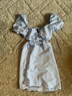 Blåvit rutig kortklänning från Divided - Supersöt blåvit rutig kortklänning från Divided i storlek 34. Klänningen har puffärmar, snörning framtill och en smockad rygg. Tillverkad i lätt bomullsmaterial, perfekt för varma dagar. Passar dig som gillar romantisk och trendig stil.