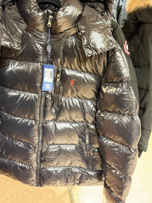 Svart pufferjacka från Polo Ralph Lauren - Snygg svart pufferjacka från Polo Ralph Lauren med glansig finish och röd logga på bröstet. Jackan har huva, flera dragkedjeförsedda fickor och quiltad design. Perfekt för kalla dagar och ger en riktigt modern look.