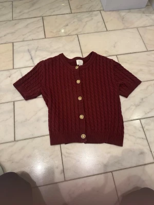 Vinröd kortärmad kofta med knappar - Säljer en vinröd kortärmad kofta med snygg kabelstickad struktur och guldfärgade knappar framtill. Tröjan har rund halsringning och ribbade kanter för en clean look. Perfekt att styla över ett linne eller t-shirt.