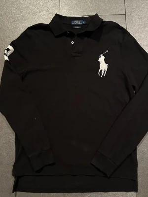 Svart långärmad pikétröja från Polo Ralph Lauren - Svart långärmad tröja från Polo Ralph Lauren i bomull med klassisk krage, vita knappar och stor vit broderad polospelare på bröstet. Snygg detalj med en vit broderad siffra 3 på ärmen. Custom fit-modell som ger en clean och sportig look.