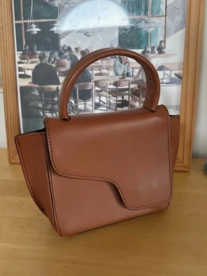 Brun handväska från ATP Atelier - Snygg brun handväska från ATP Atelier i slätt skinn. Väskan har ett stilrent och modernt formspråk med rundat handtag och magnetlås. Perfekt storlek för det viktigaste och med detaljer i guld. Märkt med ATP Atelier på insidan. Saknar det långa bandet 