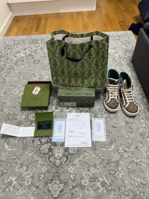 Gucci Tennis 1977 high-top sneakers - Säljer ett par Gucci Tennis 1977 high-top sneakers i beige canvas med klassiskt GG-monogram, vita detaljer och grönt innerfoder. Skorna har vita snören, röd sula med GG-mönster och gröna/röda ränder på sidan. Kommer med originalkartong, dustbag och certifikat.