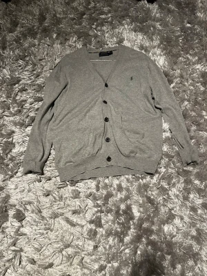 Grå kofta från Polo Ralph Lauren - Snygg grå kofta från Polo Ralph Lauren i mjuk bomull. Klassisk design med v-ringning, svarta knappar framtill och två fickor. Riktigt fet tröja nu till vår o sommar. Perfekt till lager på lager och enkel att matcha.