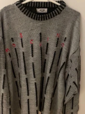 Grå stickad tröja Moschino Jeans - Säljer en grå stickad tröja från Moschino Jeans med svarta vertikala streck och diskret röd text på bröstet. Tröjan har en svartvitrandig ribbad krage och långa ärmar. Perfekt för dig som gillar statement-plagg med unik design. Vid intresse skriv för mer info! ABS! Pris går att diskutera! 