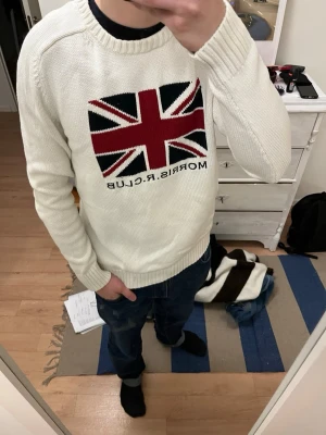 Vit stickad tröja Morris R Club - Säljer en vit stickad tröja från Morris R Club med Union Jack-flagga i rött, svart och vitt på bröstet samt texten 'MORRIS.R.CLUB' under. Tröjan har rund halsringning, ribbade muddar och lång ärm. Perfekt för dig som gillar klassisk brittisk stil. Storlek L men mer Medium i passform 