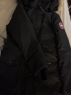  Canada Goose - Säljer en svart Canada Goose dunjacka med klassisk patch på ärmen och stora fickor framtill. Jackan har huva och stängs med både dragkedja och knappar. Perfekt för kalla vinterdagar och riktigt snygg streetstil. Säljer för 2000 men snabb affär 1500