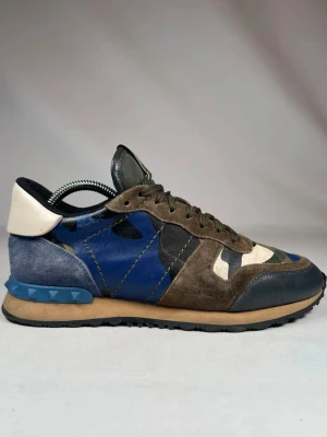 Valentino Garavani camo sneakers - Säljer ett par unika Valentino Garavani sneakers med camouflagemönster i blått, grönt, beige och svart. Skorna har detaljer i mocka och skinn, samt coola nitar på hälen. Klassisk snörning och rund tå ger en sportig vibe. Perfekta för dig som vill sticka ut med stil.