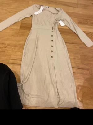 Beige omlottklänning med knappar - Snygg beige långklänning från H&M med omlott upptill och dekorativa knappar längs framsidan. Klänningen har långa ärmar, diskret ribbat mönster och markerad midja. Perfekt för dig som gillar stilrena och enkla plagg med fina detaljer.