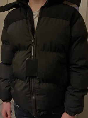 Svart pufferjacka från ByCastello - Säljer en svart pufferjacka från ByCastello med huva och dragkedja framtill. Jackan har detaljer i manchester på axlar och huva, vilket ger en schysst kontrast till det släta materialet. Perfekt för kalla dagar och riktigt snygg. Jackan har fortfarande prislappen kvar och är aldrig använd. En liten skada skedde när förpackningen skulle öppnas därav priset. Köpt för 2800kr!