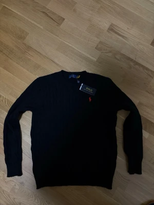 Svart kabelstickad tröja Polo Ralph Lauren - Kabelstickad svart tröja från Polo Ralph Lauren med klassisk röd logga broderad på bröstet. Rund halsringning och långa ärmar. Tillverkad i mjuk bomull som känns skön mot huden. Perfekt för dig som gillar stilrena och tidlösa plagg.