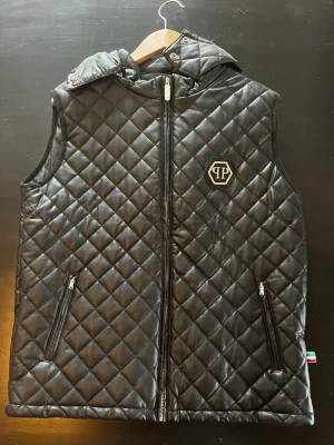Svart quiltad väst från Philipp Plein - Säljer en svart quiltad väst från Philipp Plein med avtagbar huva och dragkedja framtill. Västen har två fickor med dragkedja och ett metallmärke på bröstet. Snygg och edgy look som passar perfekt till lager-på-lager-stil.