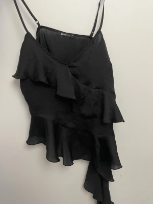 Svart volangig linnetopp Gina Tricot - Säljer en svart linnetopp från Gina Tricot med tunna axelband och asymmetrisk volangdetalj. Toppen har en v-ringad hals och är tillverkad i ett lätt, flowy material som ger en snygg siluett. Perfekt för dig som gillar statement-plagg!