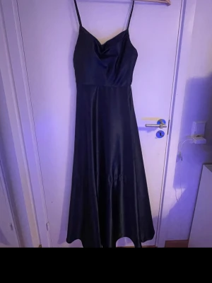 Marinblå balklänning med smala band - Säljer en elegant marinblå långklänning med smala axelband och lätt draperad urringning. Klänningen har en glansig finish och faller snyggt längs kroppen. Perfekt för dig som vill ha en stilren och klassisk look.
