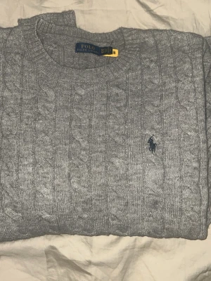 Grå kabelstickad tröja Polo Ralph Lauren - Kabelstickad grå tröja från Polo Ralph Lauren i storlek M. Klassisk rund halsringning och ikoniska broderade loggan på bröstet. Perfekt för dig som gillar stilrena och tidlösa plagg. Mjuk och skön känsla, passar till allt.