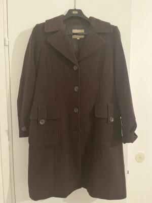 Mörkbrun ullkappa med stora fickor - Säljer en mörkbrun kappa i ull med stora fickor framtill och snygga knappar. Klassisk krage, knäppning fram och dekorativ knappdetalj bak. Perfekt för dig som vill ha en stilren och tidlös look under höst och vinter.