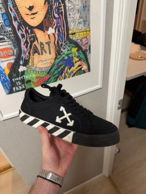 Off-White svarta sneakers med ränder - Säljer ett par svarta Off-White sneakers med ikoniska vita pilar på sidan och svartvita ränder på sulan. Skorna har snörning, platt sula och en orange detalj på sidan. Tillverkade i canvas och gummisula. Kommer med originalkartong och papper.