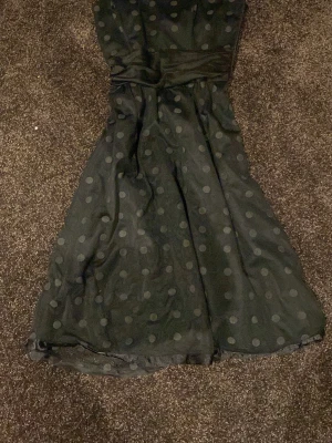 Svart prickig axelbandslös klänning - Säljer en svart axelbandslös klänning med stora prickar och markerad midja. Klänningen har en glansig finish och ett lager av tunnare tyg ovanpå. Dragkedja i sidan för enkel påklädning. Perfekt för dig som gillar klassiska detaljer och en feminin siluett.