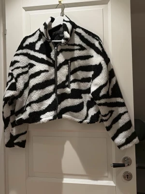 Zebramönstrad teddyjacka från Chiquelle - Säljer en fluffig teddyjacka från Chiquelle med svartvitt zebramönster. Jackan har hög krage, dragkedja framtill och en croppad passform. Perfekt för dig som vill sticka ut med en mysig och trendig look.