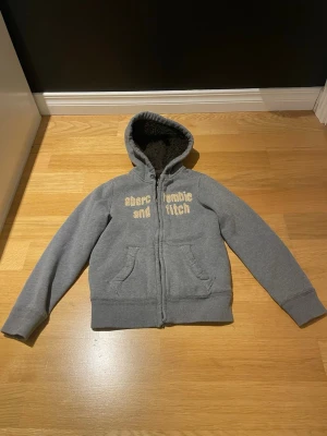 Grå hoodie från Abercrombie & Fitch - Mysig grå hoodie från Abercrombie & Fitch med broderad text på bröstet. Tröjan har dragkedja, två fickor fram och en fluffig, mörkbrun fuskpälsfodrad huva. Perfekt för kyliga dagar och riktigt skön att dra på sig.