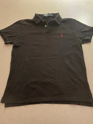 Svart piké från Polo Ralph Lauren - Klassisk svart pikétröja från Polo Ralph Lauren i slim fit. Tröjan har korta ärmar, krage med två knappar och den ikoniska röda loggan broderad på bröstet. Tillverkad i mjuk bomull som känns skön mot huden.