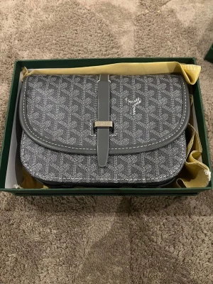 Grå axelväska  - Säljer en grå axelväska med ikoniskt geometriskt mönster och vit logga på framsidan. Väskan har ett lock med spänne och detaljer i skinn. Perfekt storlek för det viktigaste och en riktigt snygg accessoar.