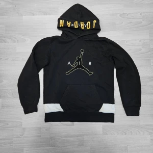 Jordan Black Hoodie - Klassisk Air Jordan hoodie i svart med fet guld broderad "AIR" Jumpman-logo fram och stor guld "JORDAN"-text runt huvan – premium streetwear-känsla. Mjuk, tjock fleece-tyg. Passar som S