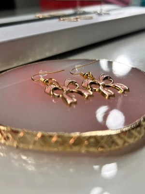 Reséglans🍸 - Säljer ett par söta örhängen i form av rosetter. De är i guldmetall med ljusrosa detaljer och har krokar för enkel användning. Perfekta för dig som vill ha en gullig och trendig accessoar till din outfit✨✨✨✨