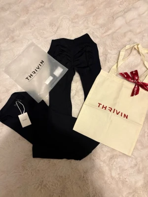 Svarta bootcut byxor från Thrivin - Säljer ett par svarta bootcut byxor från Thrivin. Byxorna har låg/mellan midja och är tillverkade i ett stretchigt material som sitter snyggt. De har en scrunch-detalj bak och vida ben. Perfekta till gymmet!