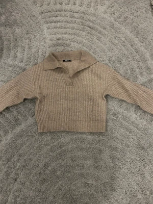 Beige stickad tröja från Gina Tricot - Säljer en beige stickad tröja från Gina Tricot med krage och v-ringad öppning. Tröjan har lång ärm och ribbade muddar samt en croppad passform. Perfekt för lager på lager under höst och vinter.