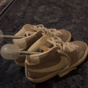 Nike Blazer Mid i beige mocka - Säljer ett par Nike Blazer Mid sneakers i beige mocka med vita detaljer och snörning. Skorna har en klassisk retrolook med högt skaft och vit sula. Perfekta för dig som gillar streetwear och vill ha en tidlös sneaker i garderoben. Storlek 36