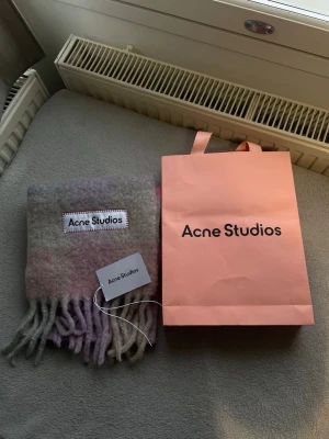 Rutig ullhalsduk Acne Studios - Säljer en fluffig och lyxig halsduk från Acne Studios i rutigt mönster med mjuka toner av grått, rosa och lila. Halsduken har fransar i ändarna och stor Acne Studios-logga. Perfekt accessoar för att höja din vinterstil.