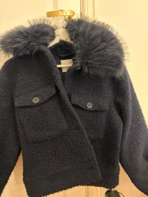 Mörkblå teddyjacka från Gina Tricot - Säljer en mörkblå teddyjacka från Gina Tricot med fluffig fuskpäls på kragen och två stora bröstfickor med knappar. Jackan har en mysig och trendig look, perfekt för kalla dagar. Passar dig som gillar en lite oversized stil.