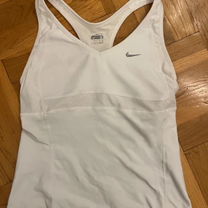 Vit sportig linnetopp från Nike - Säljer ett vitt träningslinne från Nike. Toppen har inbyggd bh och är tillverkad i ett stretchigt syntetmaterial som passar perfekt för träning. Diskret Nike-logga framtill och smickrande v-ringning.