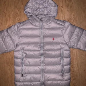 Ljusgrå dunjacka från Polo Ralph Lauren - Snygg ljusgrå dunjacka från Polo Ralph Lauren med huva och klassisk röd logga på bröstet. Jackan har två stora fickor med dragkedja och quiltad design. Perfekt för kalla dagar och riktigt skön att bära.