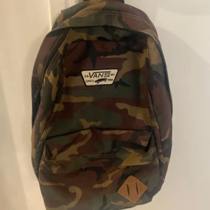Vans Camouflage ryggsäck   - Säljer en klassisk ryggsäck från Vans i camouflage-mönster med gröna, bruna och svarta toner. Väskan har ett stort huvudfack, innerficka med dragkedja och en mindre ytterficka. Tillverkad i slitstark polyester och prydd med Vans-logga framtill.