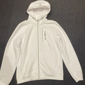 Vit hoodie från Sail Racing med dragkedja storlek 176 - Vit hoodie från Sail Racing med hel dragkedja framtill, bröstficka med dragkedja och logga på bröstet. Tröjan har huva med snörning och långa ärmar. Perfekt för dig som gillar stilren och sportig look.