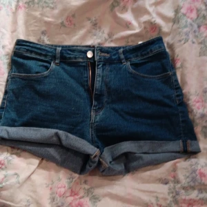 Blå jeansshorts från &Denim, strl 40 - Säljer ett par blå jeansshorts från H&M i storlek 40. Modellen är midwaist med stretch, klassisk femficksdesign och uppvikta benslut. Shortsens material är jeans och de har dragkedja och knapp framtill. Perfekta för sommaren!