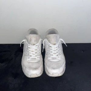 Vita Nike Air Max 90 - Säljer ett par klassiska vita Nike Air Max sneakers med mesh och skinnpaneler. Skorna har en chunky sula med synlig Air-enhet och rund tå. Perfekta för dig som gillar en clean och sportig look. Snörning framtill och diskret Nike-logga på sidan.