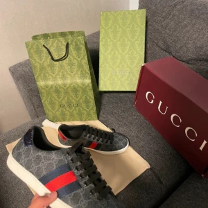Gucci sneakers med GG-mönster - Snygga Gucci sneakers i mörkgrått med klassiskt GG-mönster, svarta skosnören och vit sula. Sidan pryds av de ikoniska röd- och marinblå ränderna. Skorna har rund tå och är tillverkade i canvas med skinn-detaljer. Perfekta för dig som vill sticka ut med lyxig streetstyle.