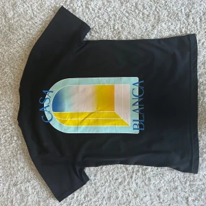 Casa Blanca  - Svart t-shirt från Casa Blanca med färgstarkt grafiskt tryck i blått, gult och vitt på både fram- och baksidan. Trycket har en cool geometrisk design och texten 'Casa Blanca' i blått. T-shirten har rund hals och korta ärmar, perfekt för en chill och stilren look.