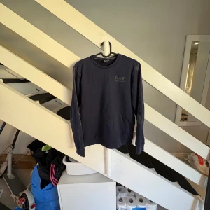 Mörkblå EA7 Emporio Armani tröja - Snygg mörkblå tröja från EA7 Emporio Armani med tryck på bröstet och ärmarna. Tröjan har rund halsringning och långa ärmar med ribbade muddar. Perfekt för dig som gillar stilrena och sportiga plagg.