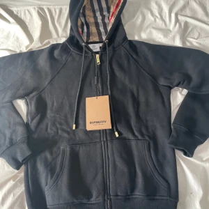 Burberry Zip Up - Svart - S - Produkt: Burberry zip-up hoodie Färg: Svart Storlek: Small Skick: Perfekt skick Detaljer: Slim fit, hel dragkedja, metalländar på snörena med “Burberry”-gravyr, dragkedja med Burberry-tryck, bekväm och stilren