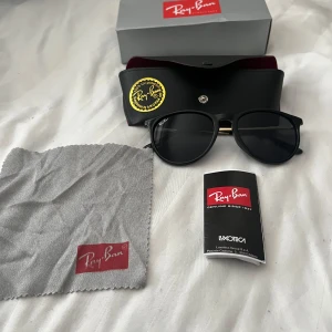 Svarta Ray-Ban solglasögon med gulddetaljer - Ray-Ban solglasögon med rundad svart båge och snygga guldfärgade skalmar. Klassisk design med mörka linser och Ray-Ban-logga på skalmen. Levereras med originalfodral, putsduk och manual. Perfekt för dig som vill ha en stilren och ikonisk look.